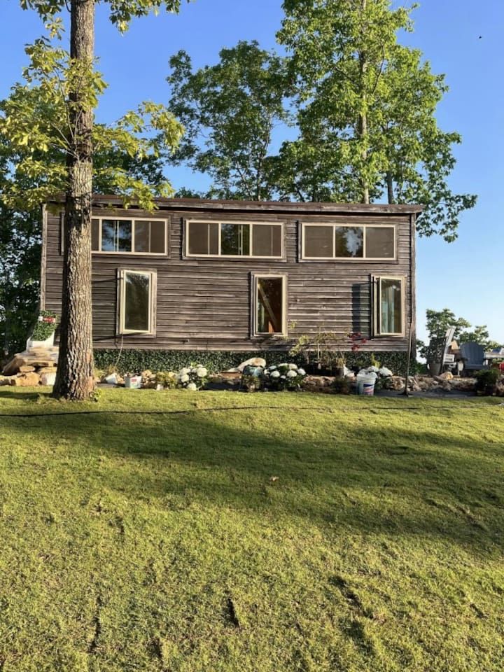 Brown wooden tiny house with multiple windows, surrounded by trees and grass.