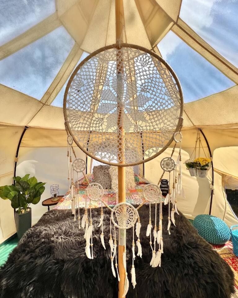 Inside a yurt, a large crochet dreamcatcher hangs. A bed with black fur blanket and pillows, and decorative accents.