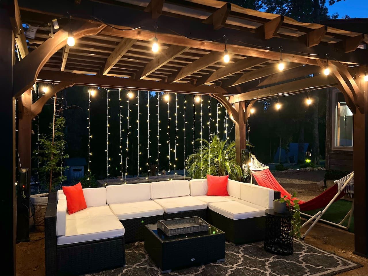 Outdoor patio with a white sectional sofa, string lights, and a fire pit under a wooden pergola at night.