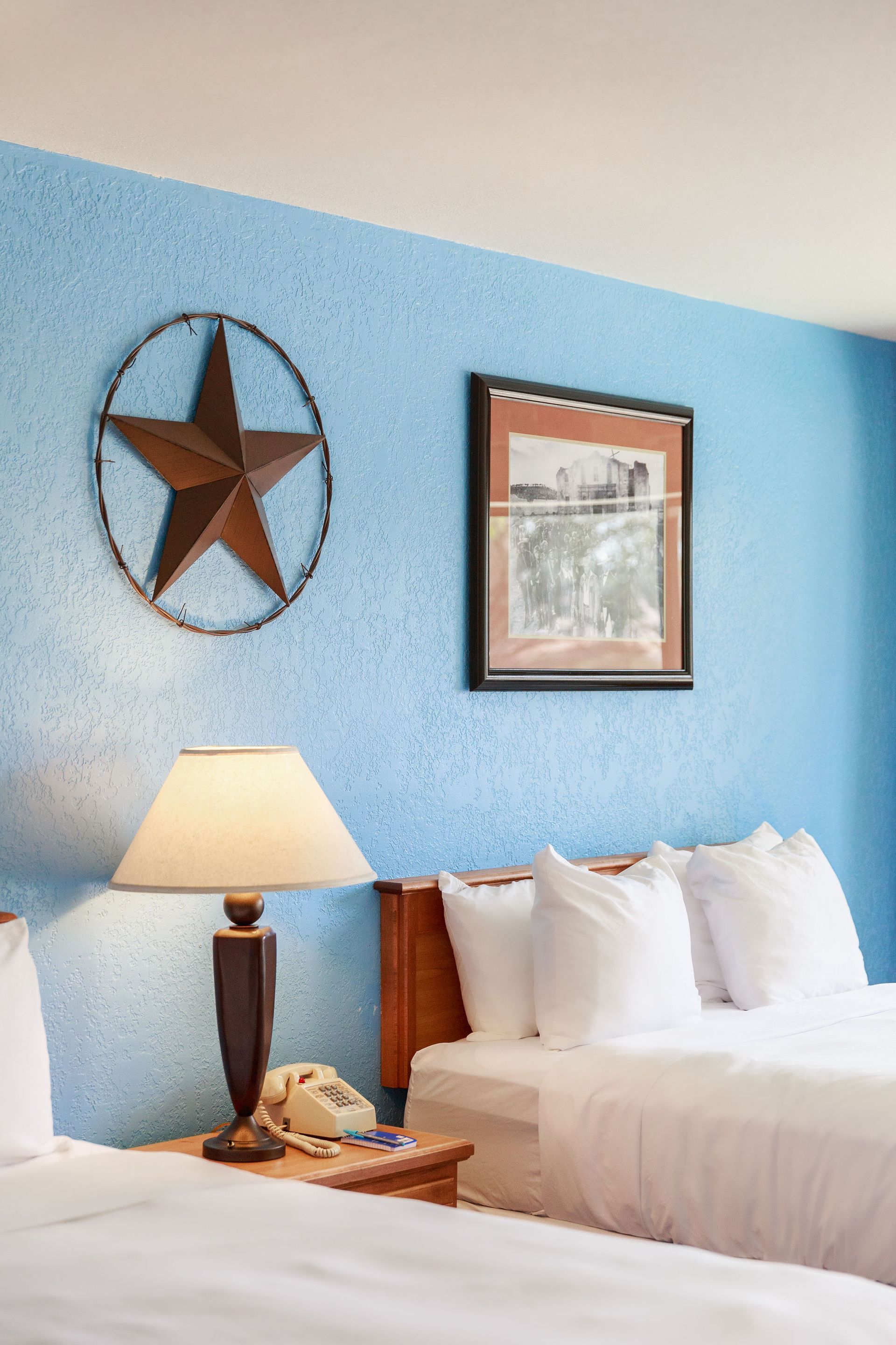 Hotel room with two beds, blue textured wall, star decor, lamp, framed pictures of John Wayne