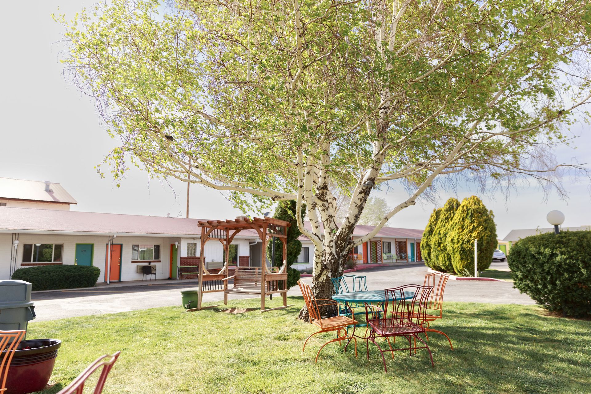 Motel exterior with tree-shaded seating area. Motel rooms in a row, brightly colored doors.