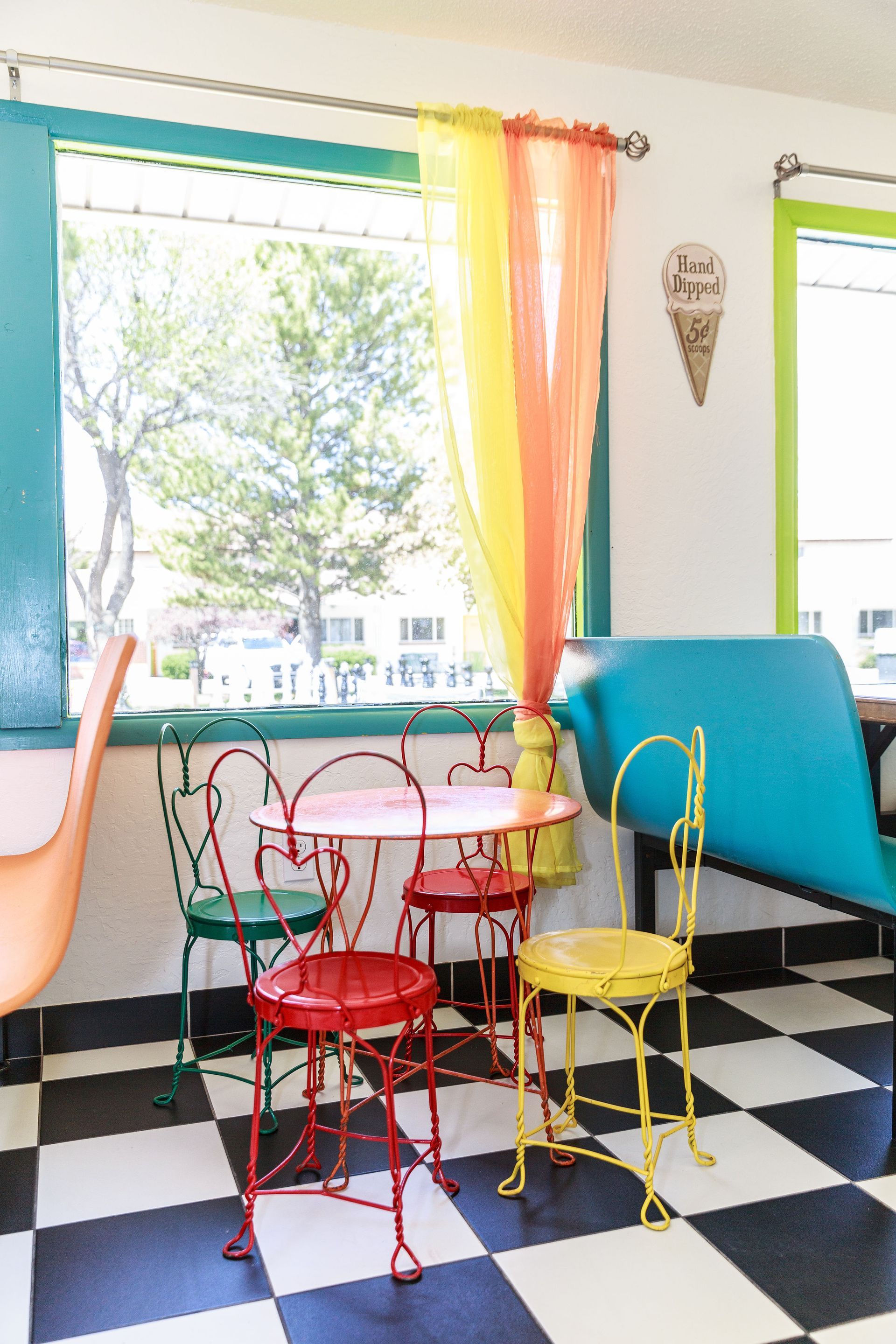 Colorful breakfast with red, yellow, and green chairs around a pink table, against a window with orange and yellow curtains.