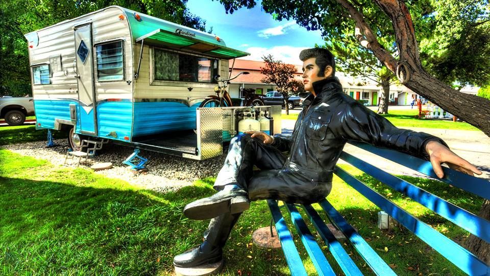 Statue of Elvis in jumpsuit sitting on a bench next to the retro inn sign and a vintage blue and white trailer.