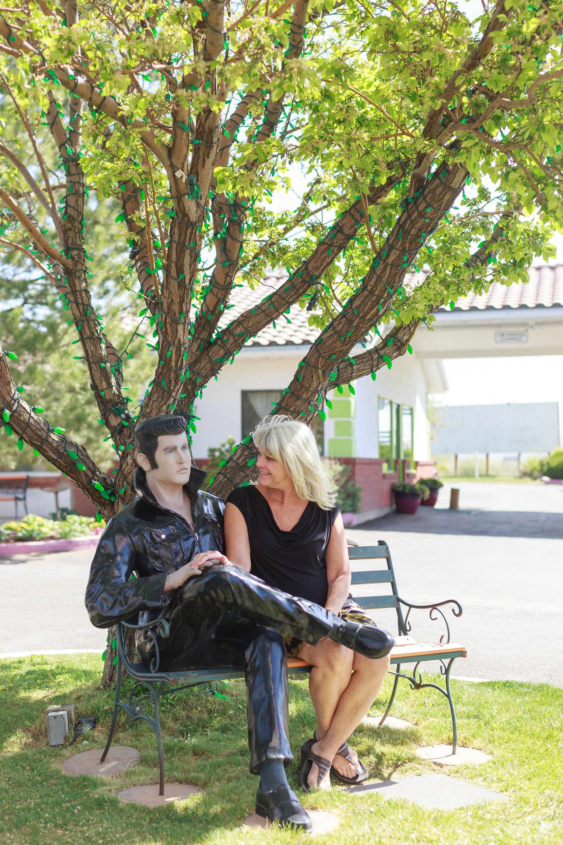 Woman sits on a bench with an Elvis statue under a tree.