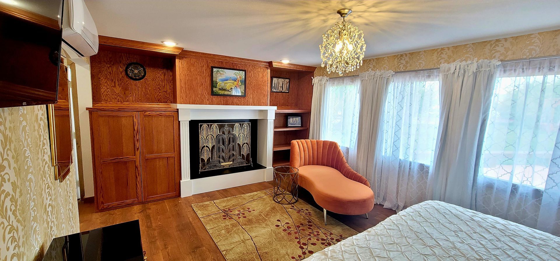 Cozy bedroom with fireplace, chaise lounge, and chandelier. Wooden cabinets and flooring, orange walls. White curtains