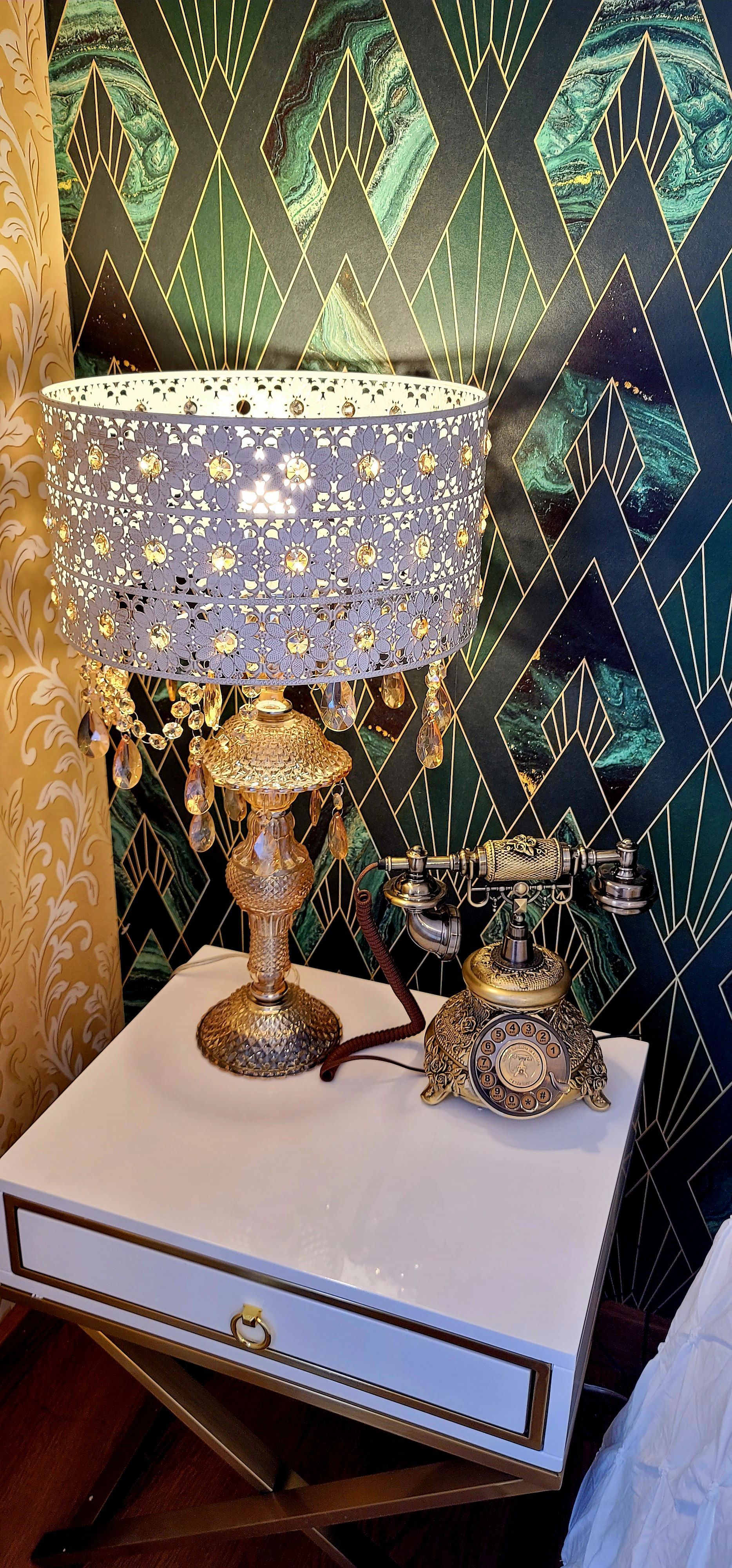 A white nightstand with a gold lamp and a vintage telephone. Dark green wallpaper in the background.
