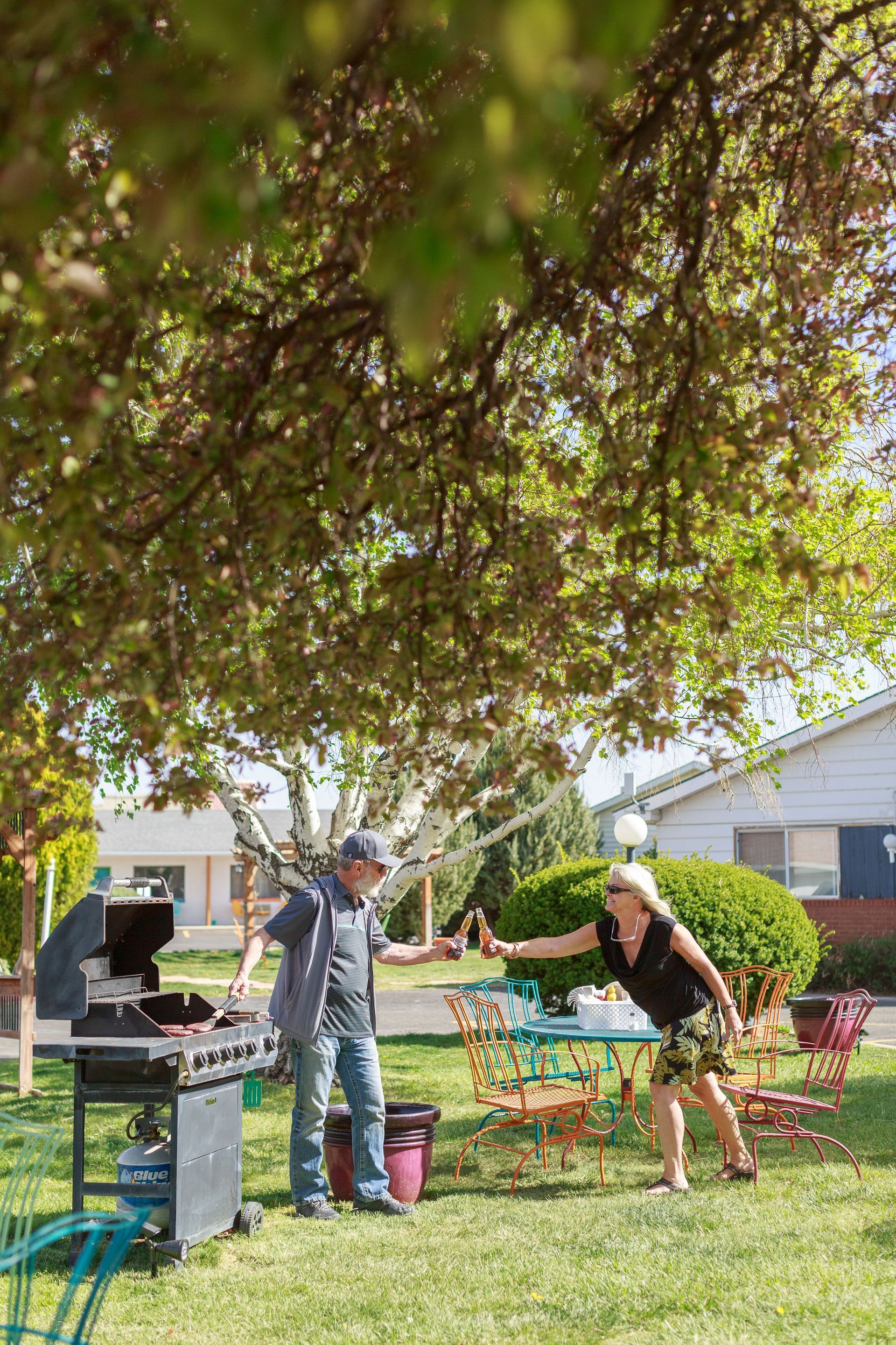 A man and woman clink bottles near a grill and table in a backyard.