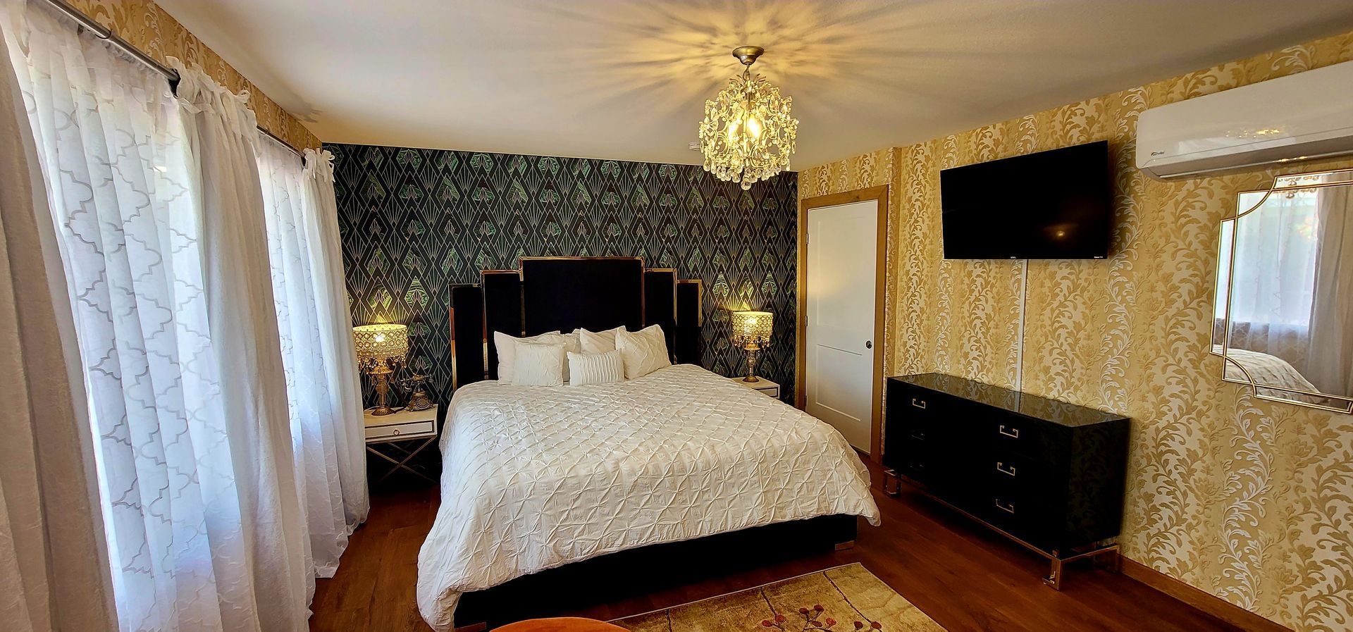 Bedroom with a bed, dresser, TV, and decorative chandelier. Wallpapered walls and sheer curtains.