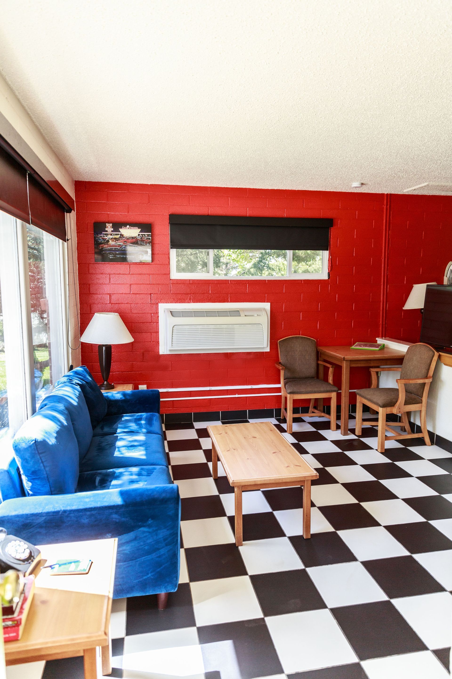 Living room: blue couch, red walls, checkerboard floor, small table, chairs, window with black blind.