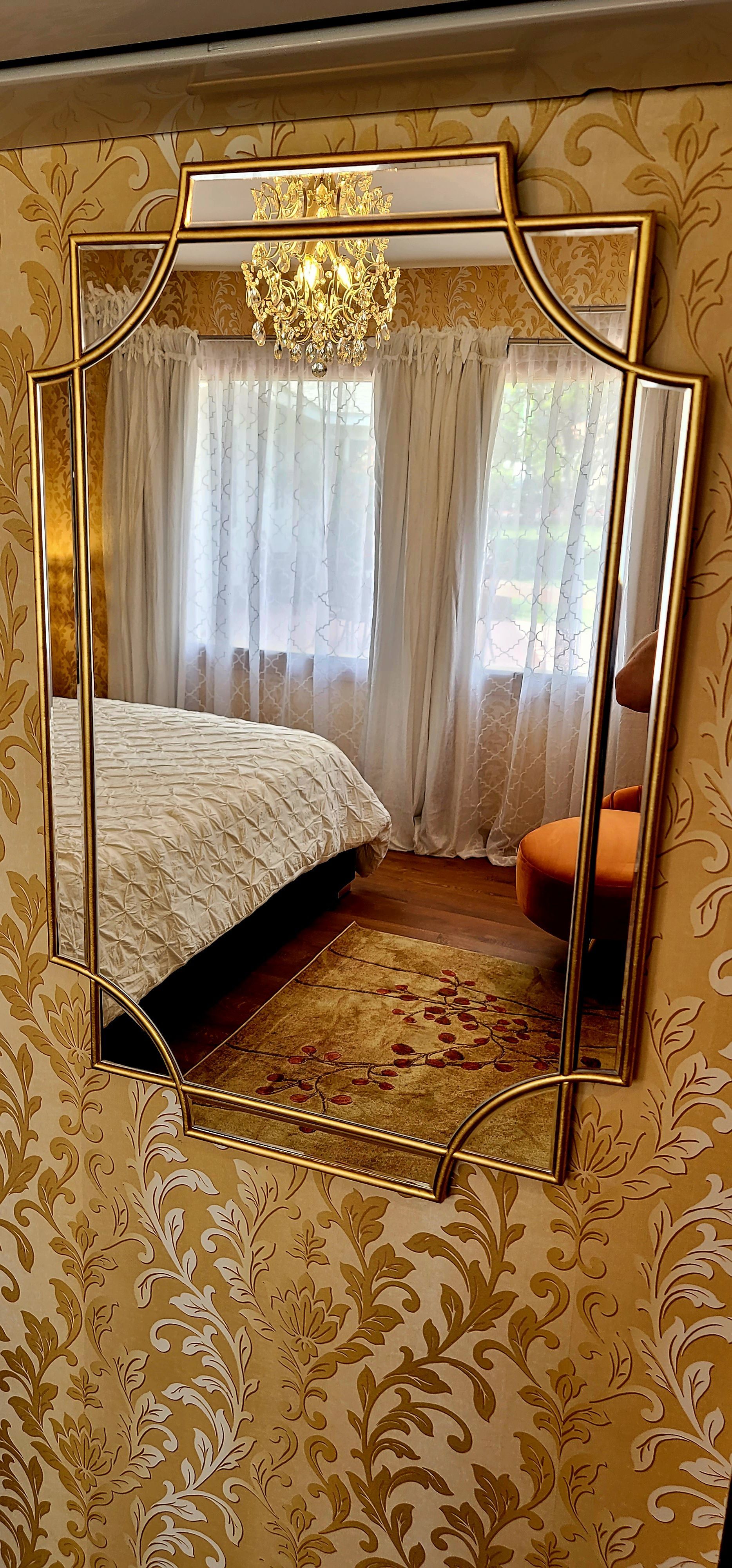 Gold-framed mirror on a floral wallpapered wall reflects a bed, rug, curtains, and an orange chair in The Great Gatsby Room.