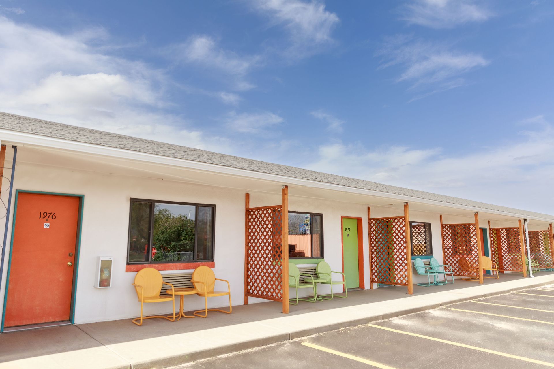 Motel exterior with rooms, orange,blue and green doors, colorful chairs, under blue sky.