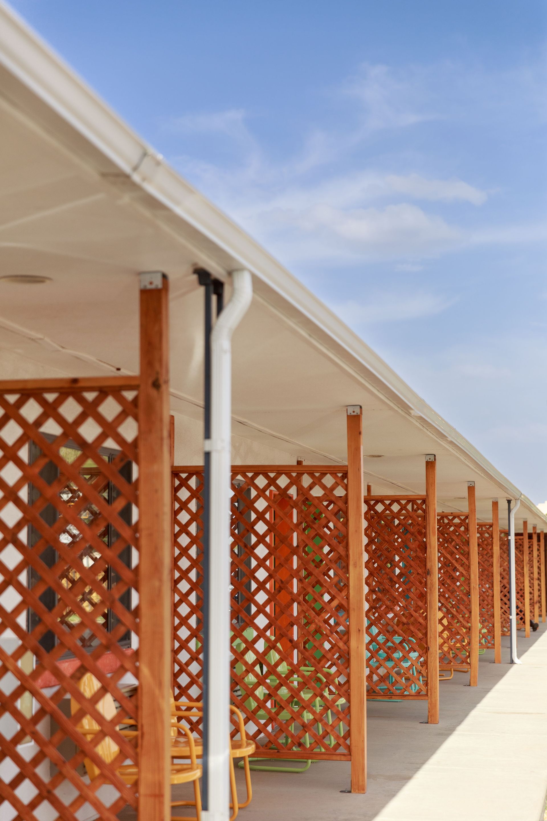 Row of motel rooms with wooden lattice walls and brown pillars, under a white awning and blue sky.