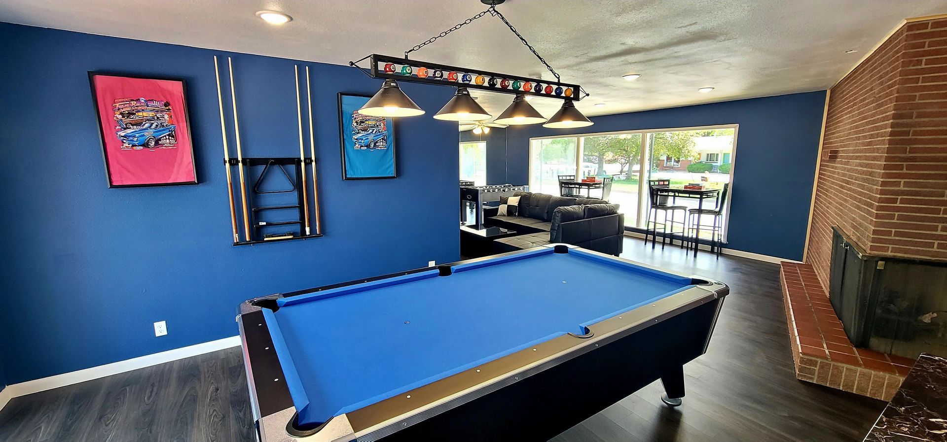 A blue-walled game room with a pool table, foosball table, videogames, leather sectional, brick fireplace, and large windows.