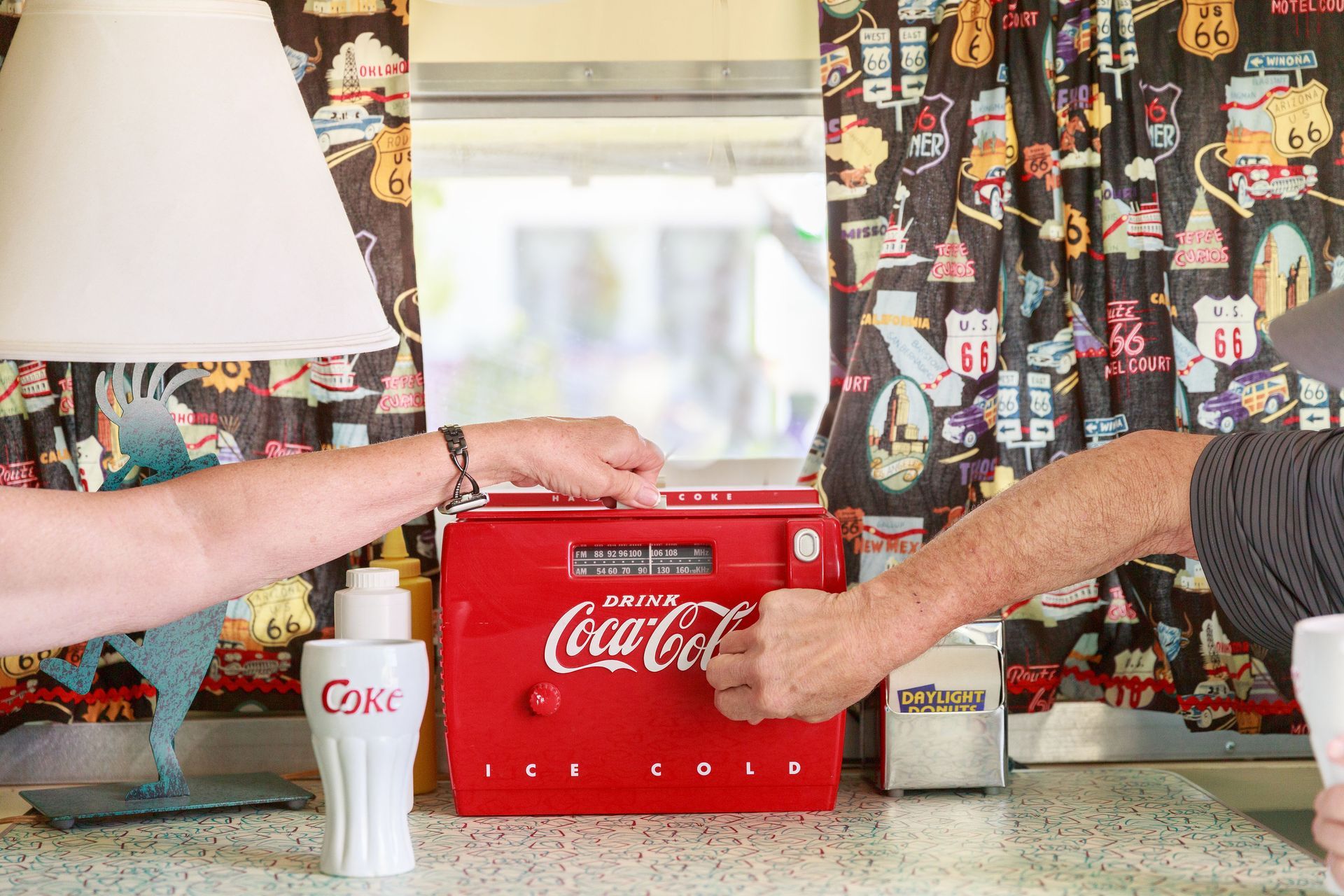 Two people turning on a vintage Coca-Cola radio in the Retro Inn vintage camper, near a window with Route 66 curtains.
