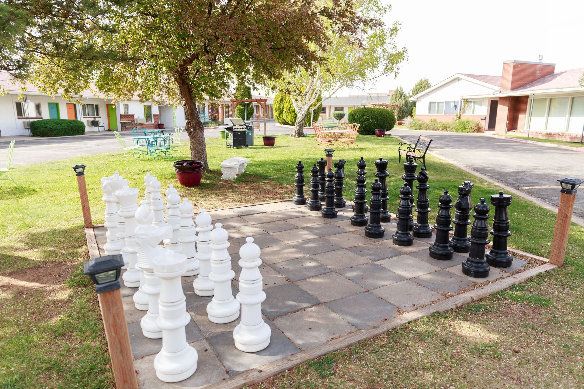 Large outdoor life sized chess set with black and white pieces on a tiled area in a grassy yard, motel in the background.