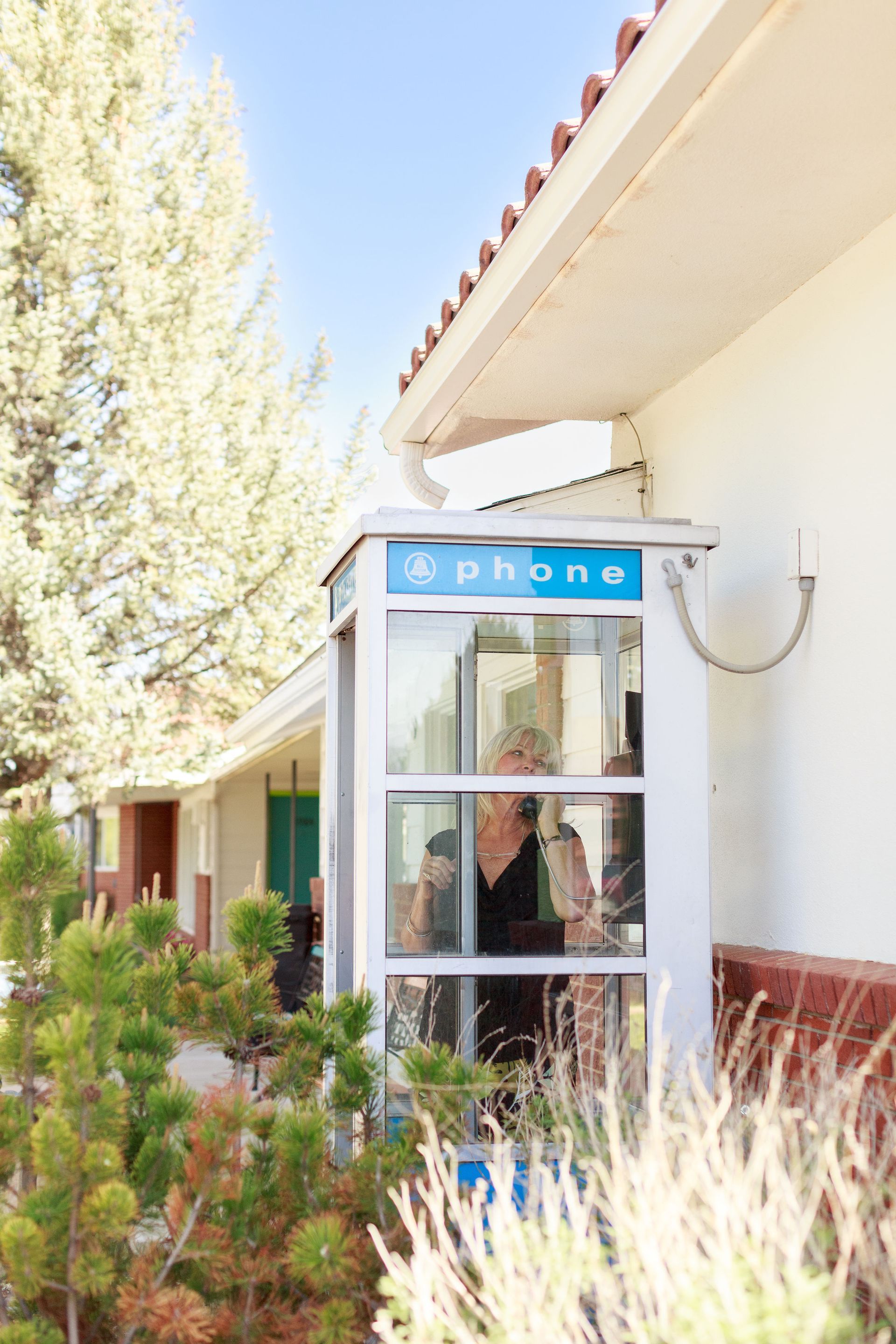 A person in a glass phone booth outside the Retro Inn. Sunlight, greenery, and blue accents.