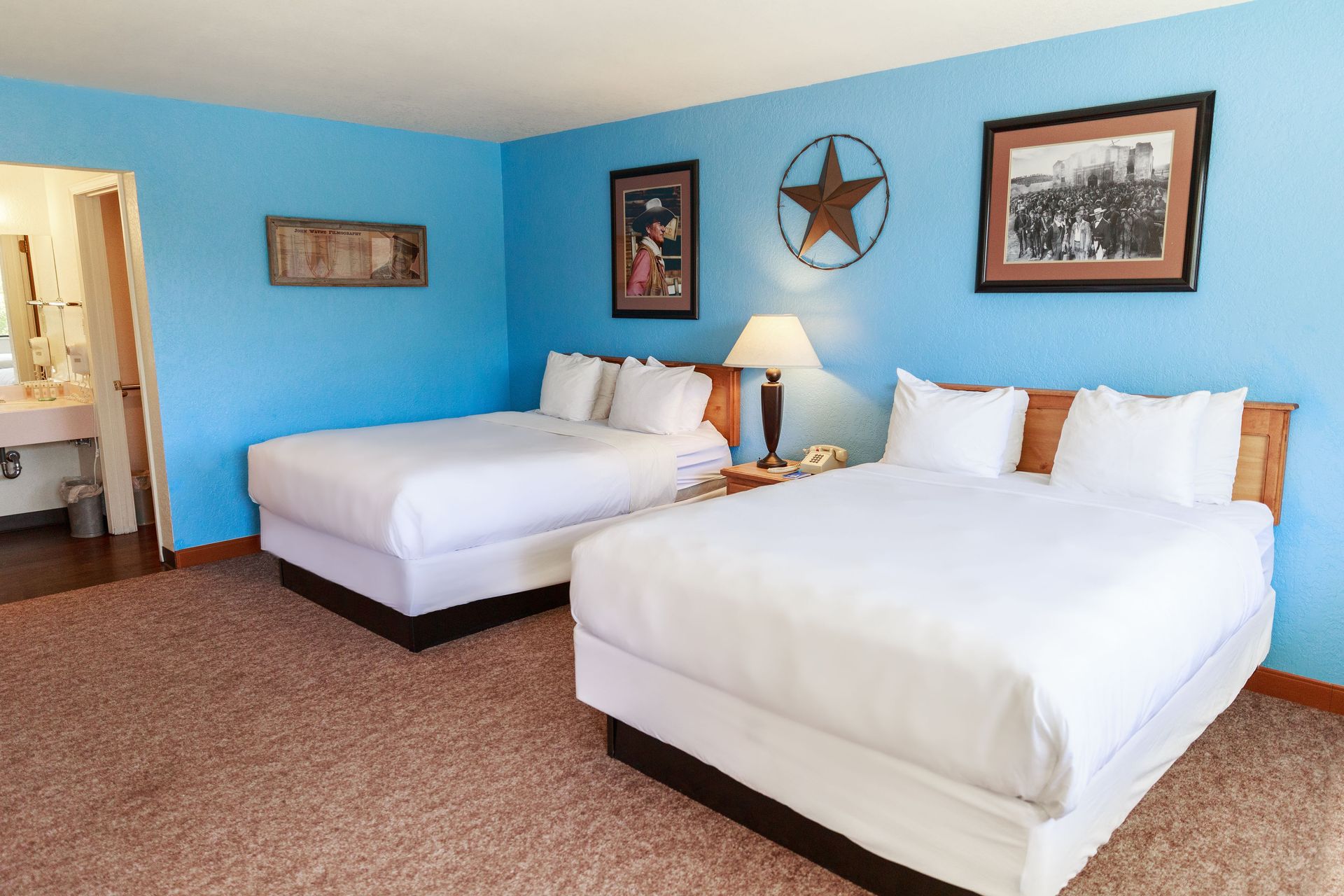 A motel room with two beds, light blue walls, John Wayne photos, and a view of a bathroom.