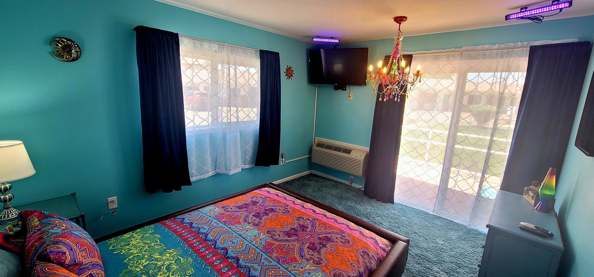 Bedroom with turquoise walls, bed with colorful cover, chandelier, TV, and sliding glass door.