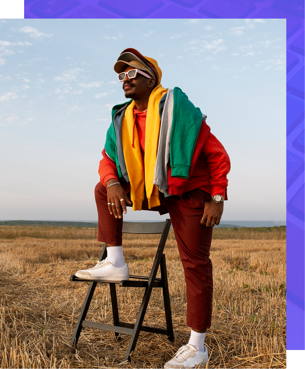 Person in colorful jacket and scarf posing on a stool in a dry field under a blue sky.