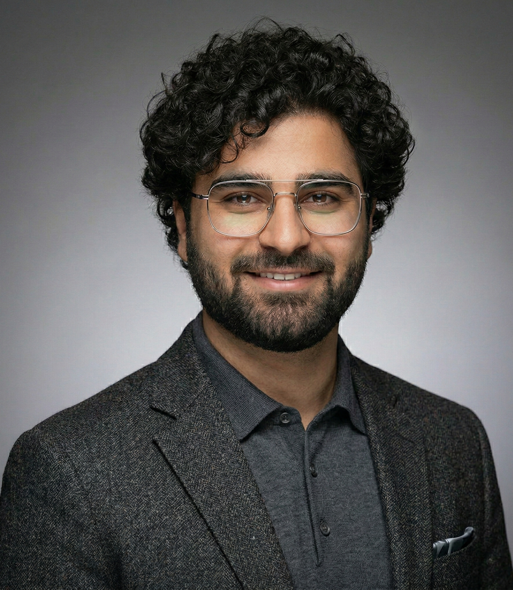 Man with curly dark hair, beard, and glasses smiles, wearing a grey jacket over a grey shirt.