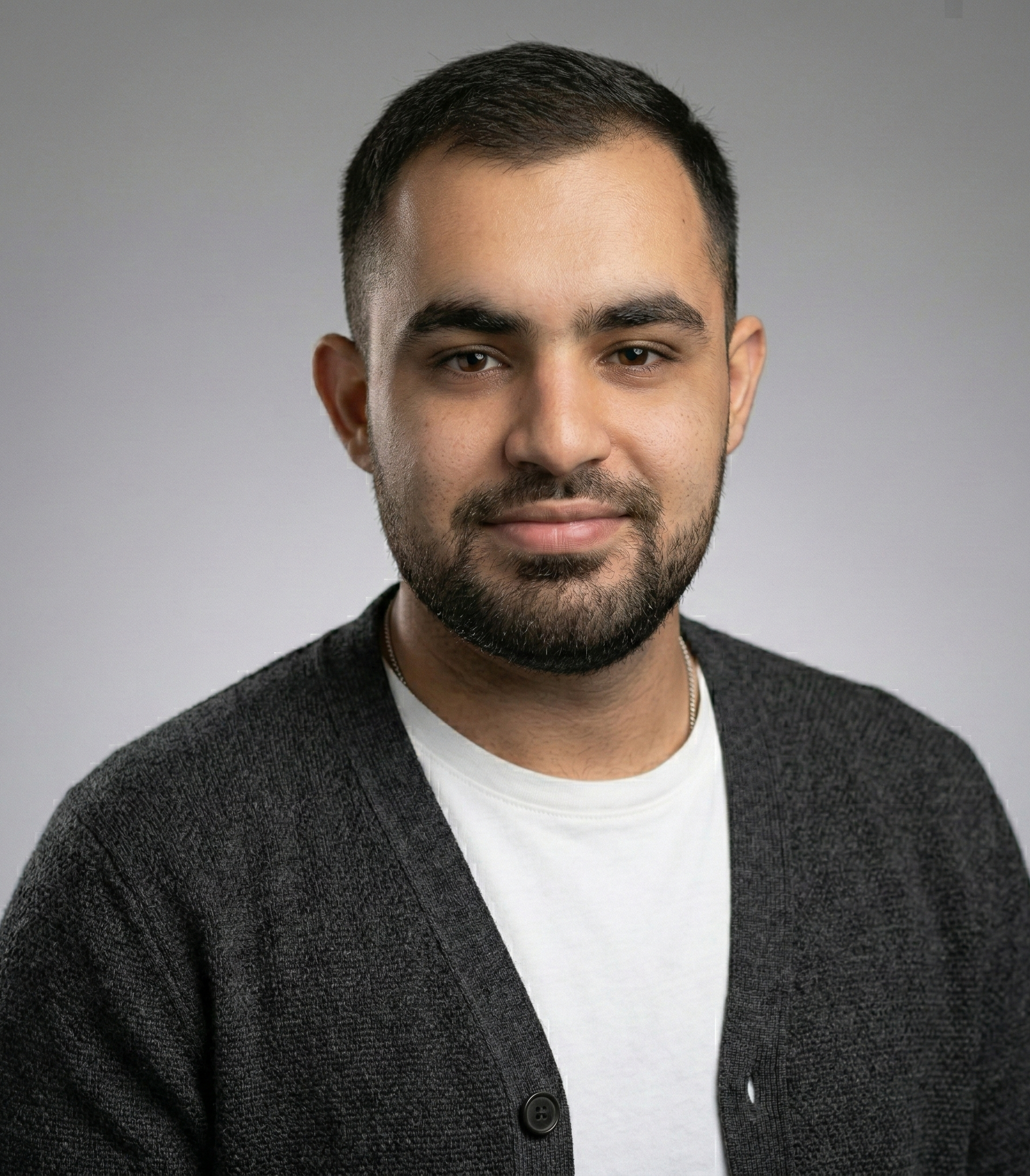 Man with short dark hair, wearing a white shirt under a gray cardigan, smiles at the camera against a gray backdrop.