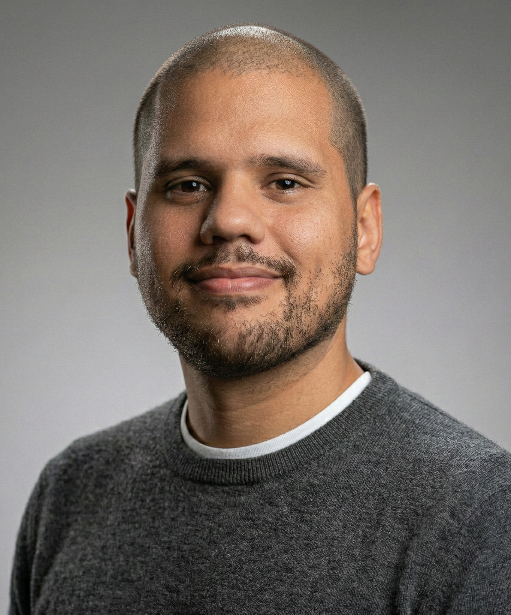 Man with a shaved head and beard, smiling, wearing a gray sweater, against a gray background.