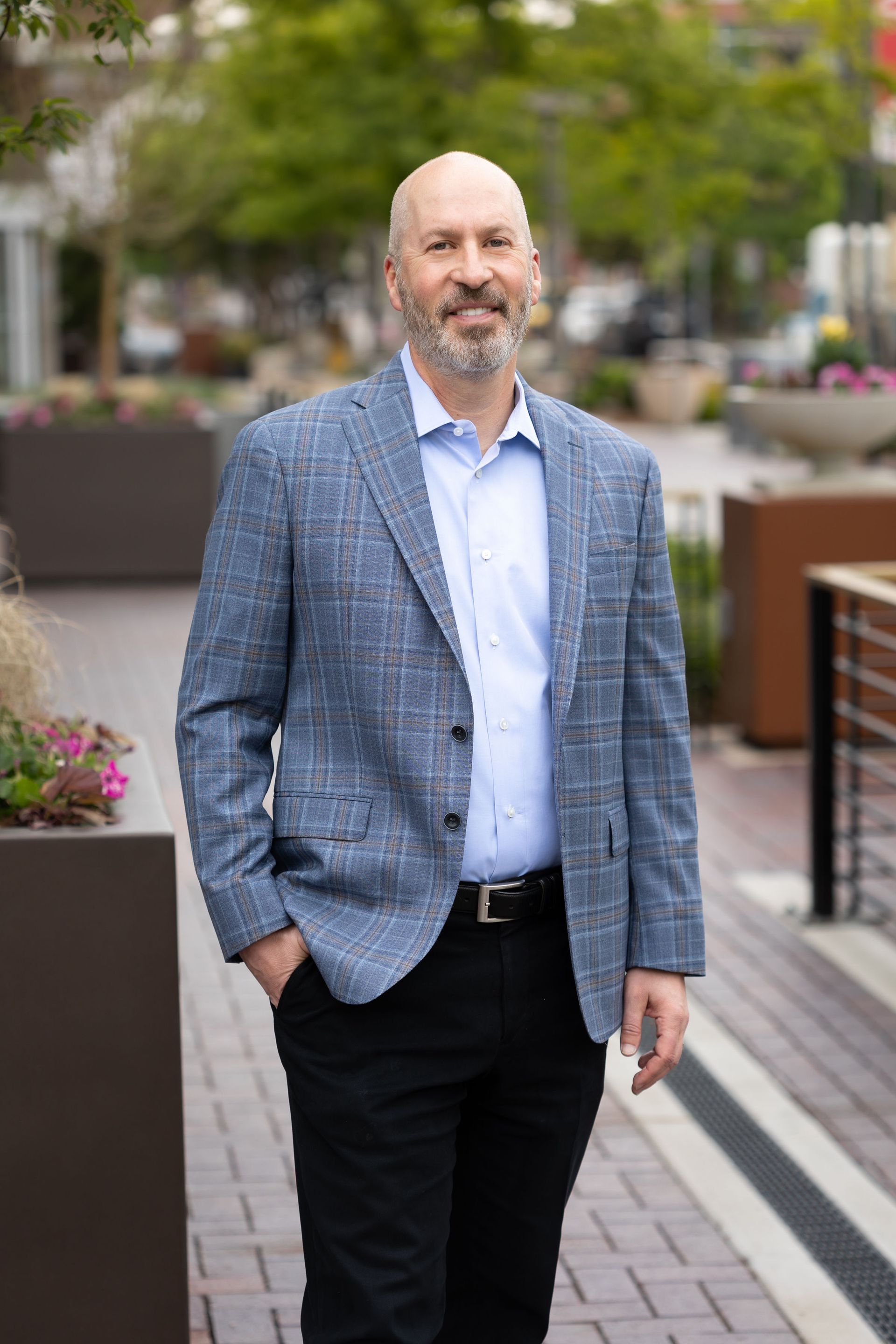 A man in a blue jacket and black pants is standing on a sidewalk with his hands in his pockets.