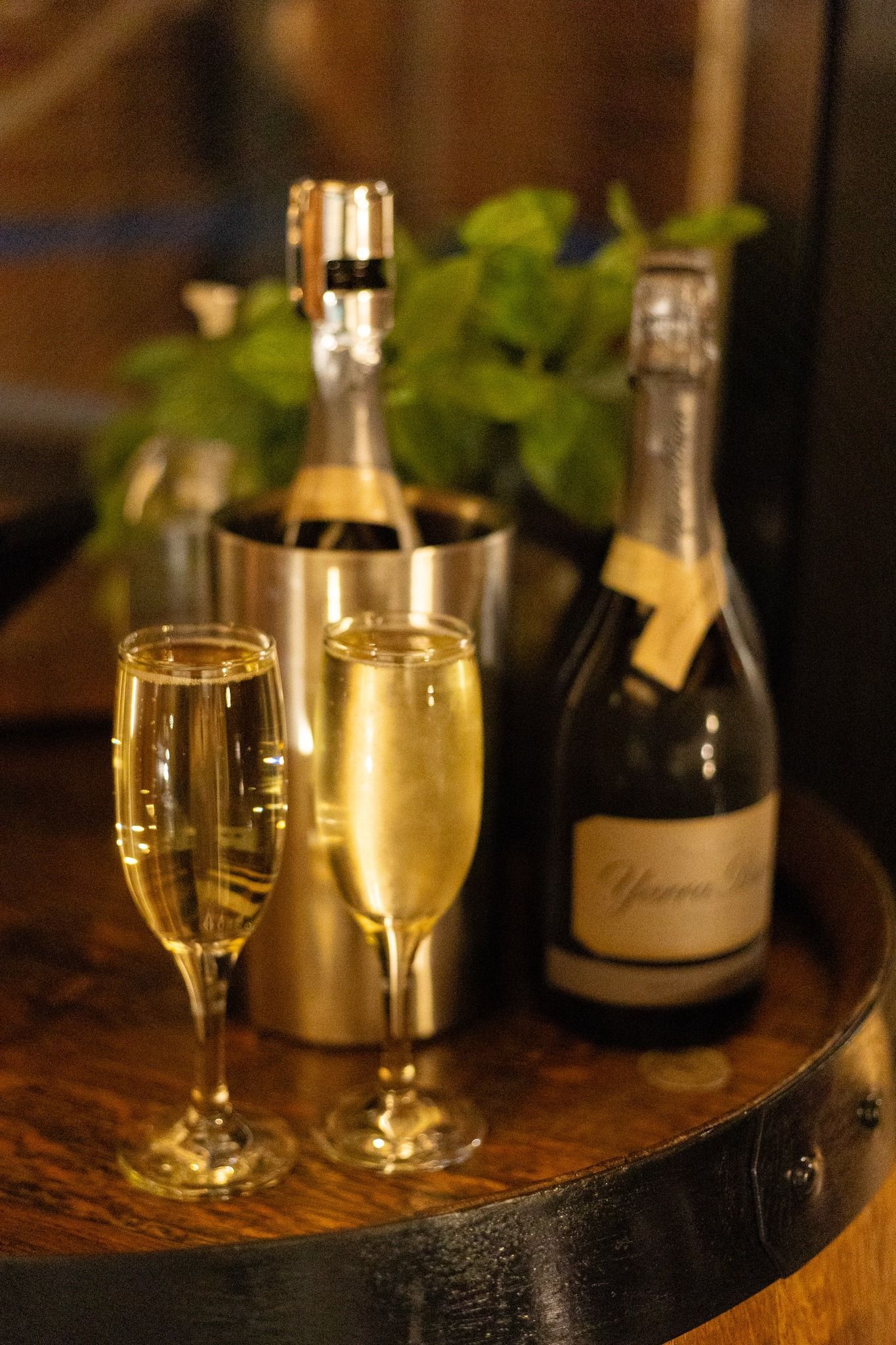 Two champagne flutes and two bottles on a wooden barrel, one bottle in a silver cooler.