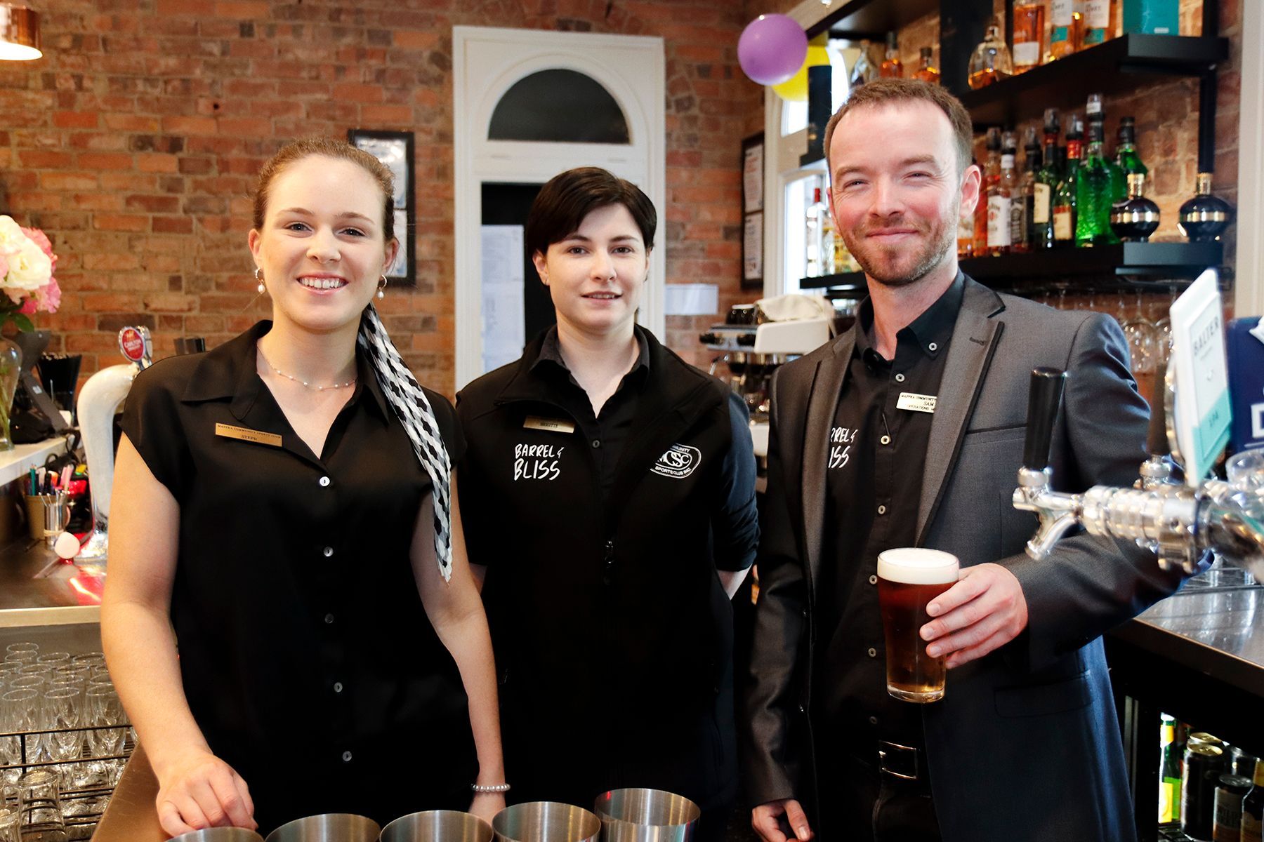 Three bar staff smiling. Two women in black shirts, a man in a jacket holding a beer at a bar.