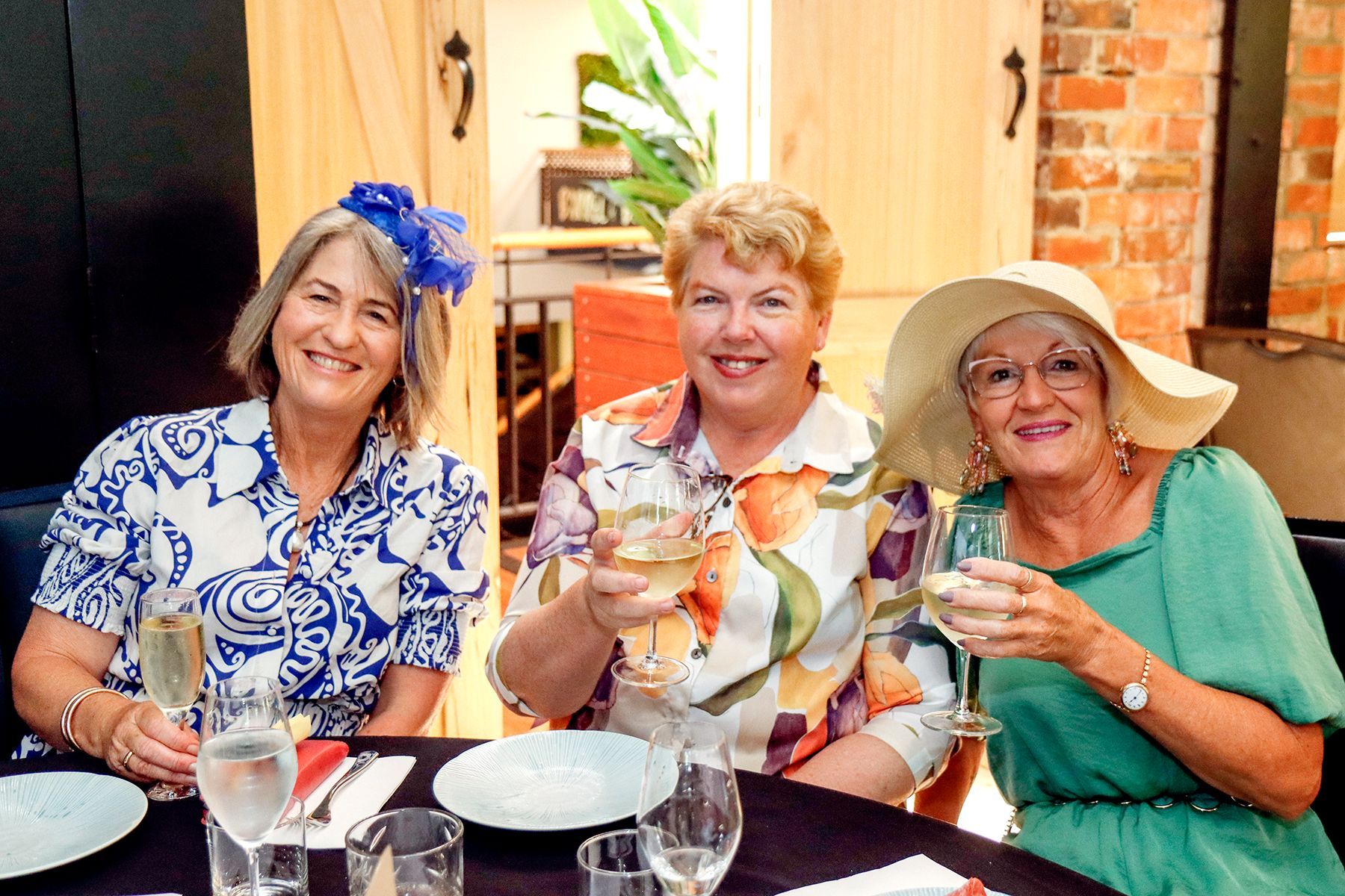 Three women in festive hats at a table, toasting with drinks. Interior with wood accents and brick.