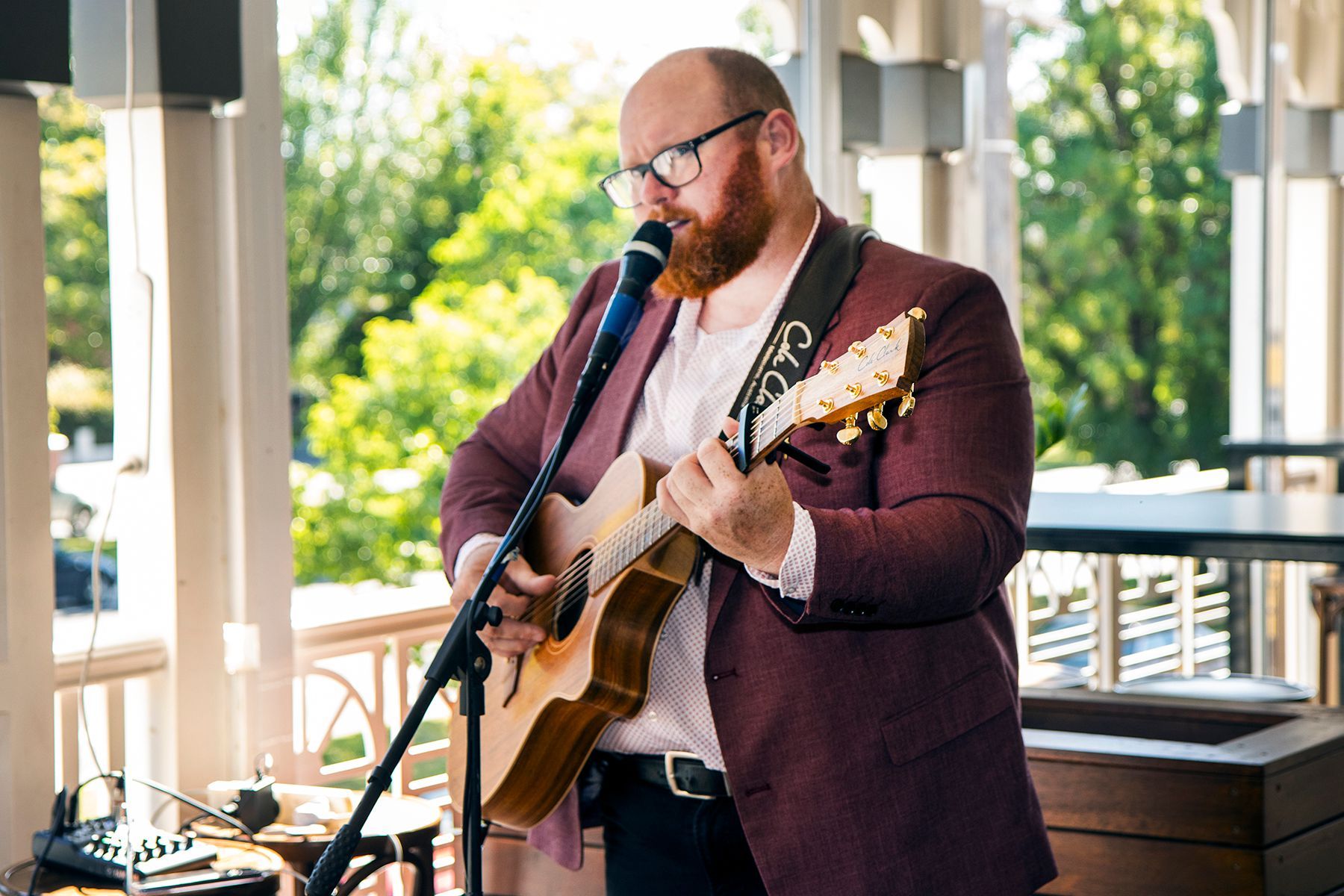Man with red beard plays acoustic guitar and sings into a microphone, wearing glasses and a red blazer, outdoors.