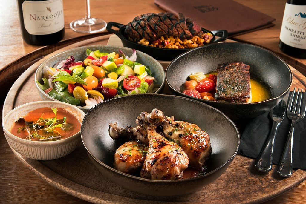 A tray with diverse dishes: chicken, salad, soup, steak and sides, wine bottles, and silverware on a table.