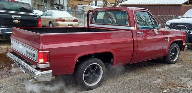 Red classic pickup truck parked on a wet lot, rear three-quarter view.