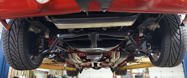Undercarriage of a red car on a lift in an auto shop, showing suspension and exhaust components.