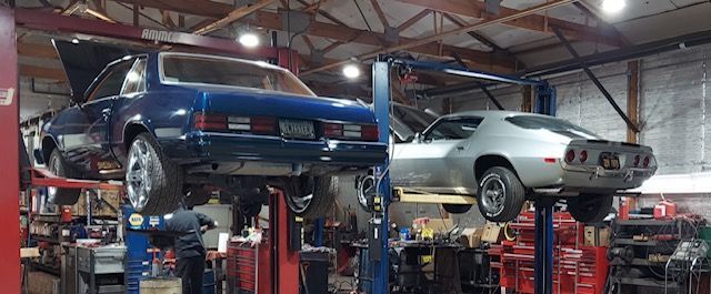 Two cars on lifts in an auto repair shop, a blue car on the left and a silver car on the right.