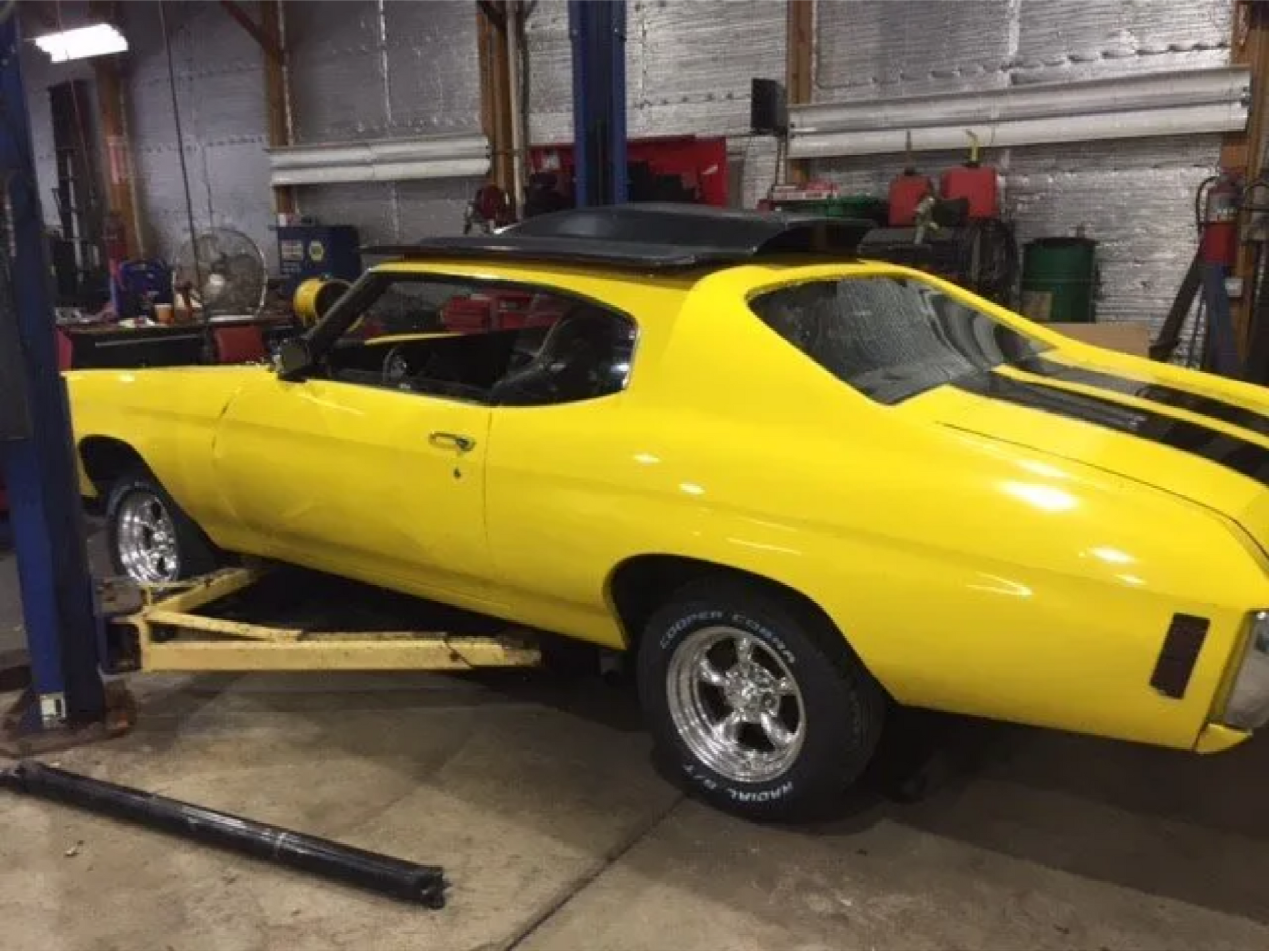 Yellow classic muscle car in a garage, lifted on a jack with tools nearby.
