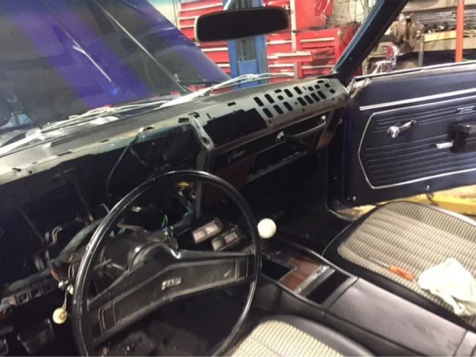 Classic car interior with black dashboard, steering wheel, and open passenger door showing a patterned seat