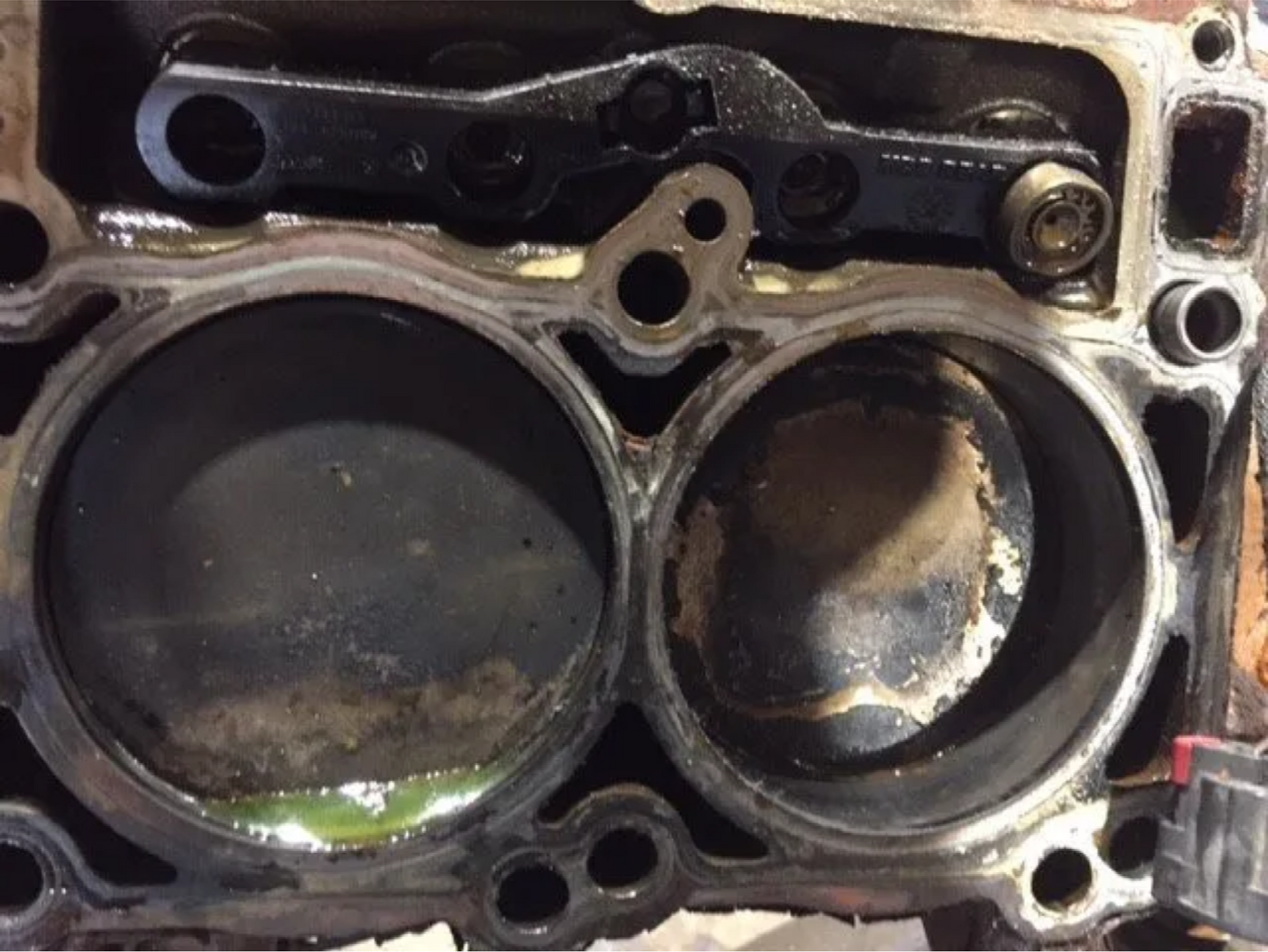 Close-up of an engine cylinder head with two piston bores, showing heavy carbon buildup and wear.