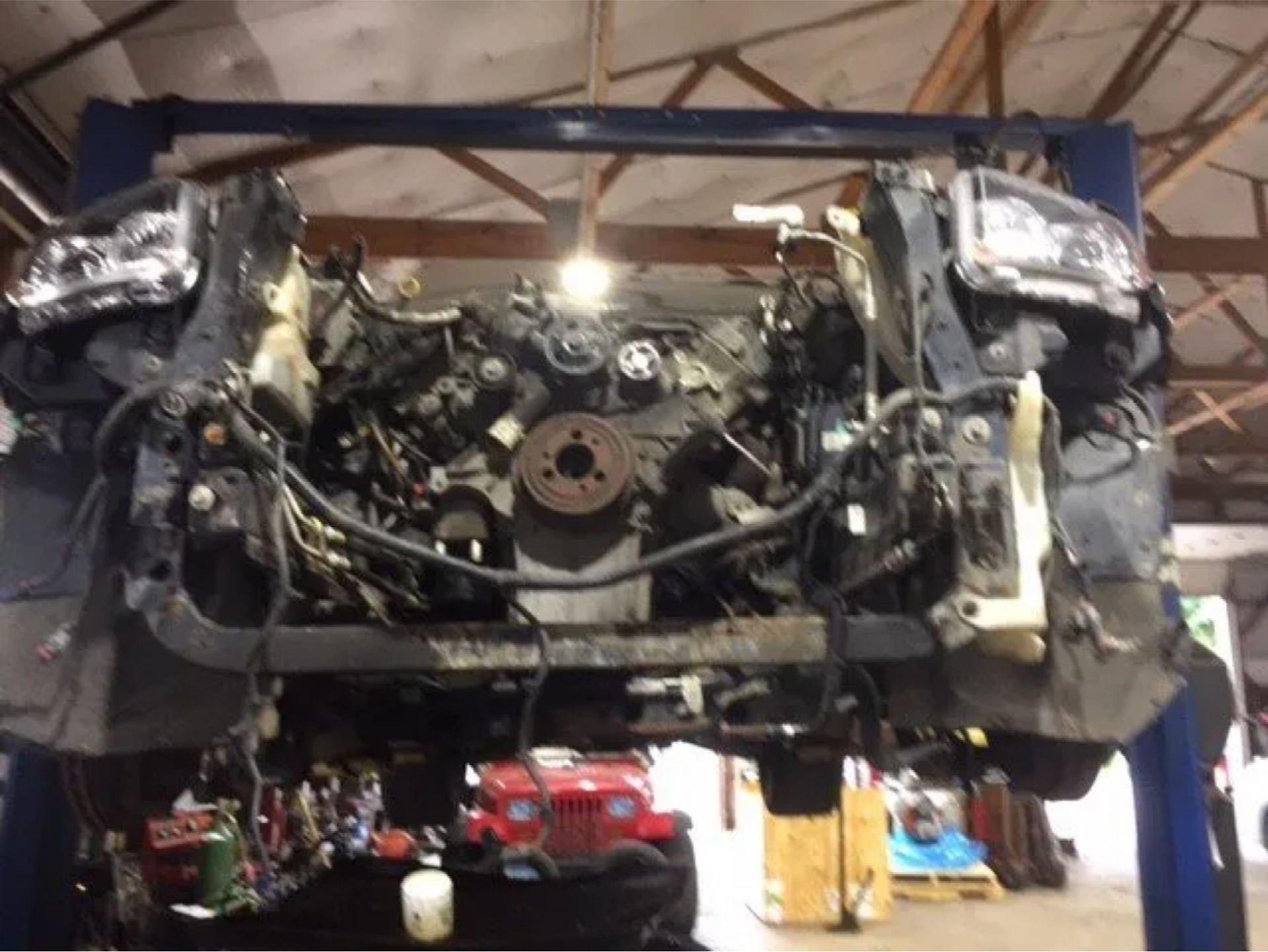Car engine bay with front body panels removed, showing exposed engine and wiring on a garage lift