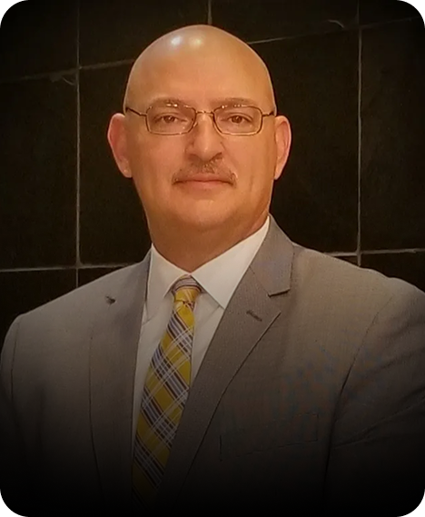 A person in a gray suit, white shirt, and yellow plaid tie, centered against a dark, tiled background.