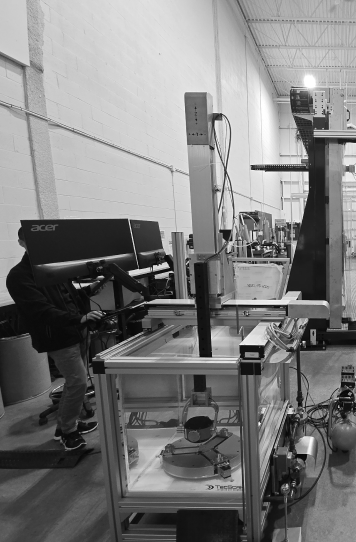 A person operates a robotic testing apparatus featuring a vertical probe assembly inside a metal frame in a warehouse.