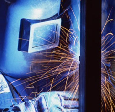 A welder in a protective mask works on metal, producing a bright shower of orange sparks.