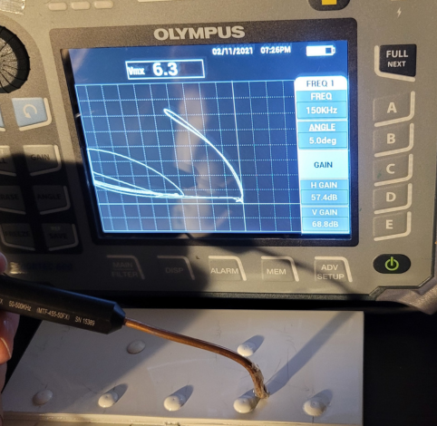 An Olympus eddy current tester displays a signal curve while a probe is used to inspect fasteners on a white surface.