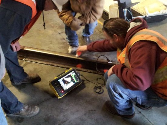 Workers in high-visibility vests inspect a rail section using a handheld ultrasonic testing device on a factory floor.