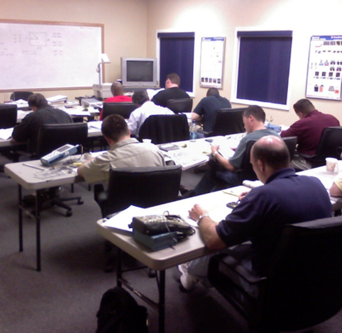 A classroom with several people sitting at folding tables, studying or working on materials.