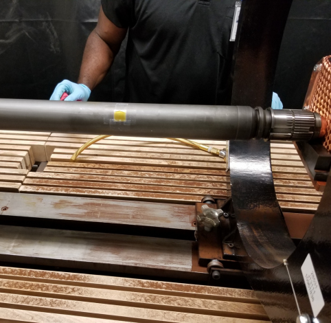 A gloved hand holds a tool near a metal cylindrical rod positioned over a wooden slat surface on industrial machinery.