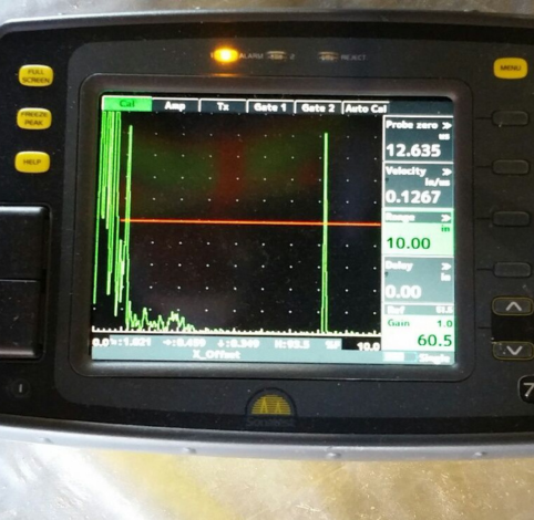 A handheld ultrasonic flaw detector showing a green signal waveform on a screen against a black background.