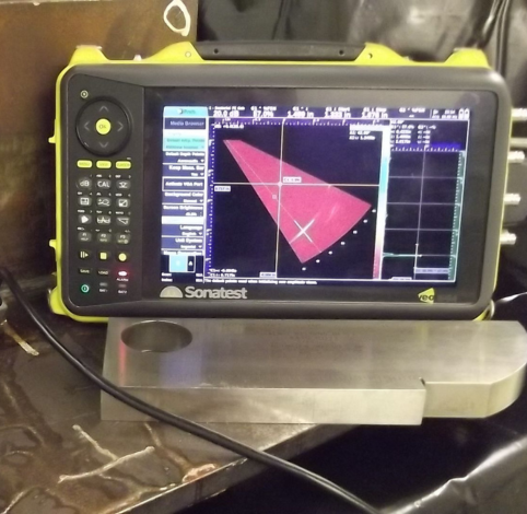 A yellow and black Sonatest ultrasonic flaw detector displays a red beam scan on its screen next to a metal test block.
