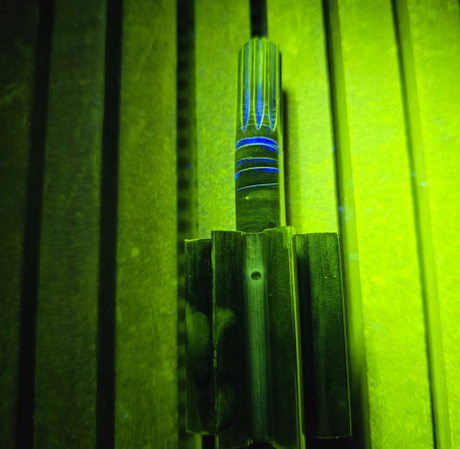 A metal object with blue fluorescent lines stands upright on a slotted green surface under ultraviolet light.