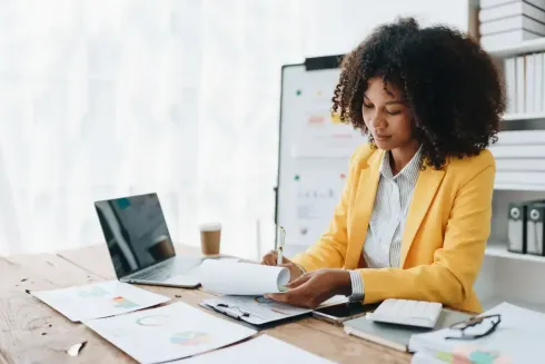 Une femme en blazer jaune examine des documents à un bureau avec un ordinateur portable dans un bureau lumineux